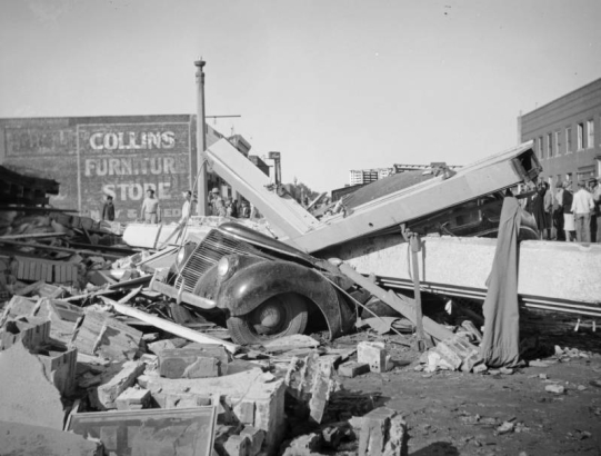 View of tornado damage to the Safeway grocery store in Julesburg (Sedgewick County), Colorado; shows a Ford sedan crushed under beams. People walk among debris; lettering reads: "Collins Furniture Store."