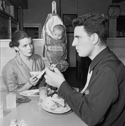 A man and woman eat sandwiches in (probably) Denver, Colorado; a baby hangs in a canvas baby carrier (cradleboard) from a hook on the wall.