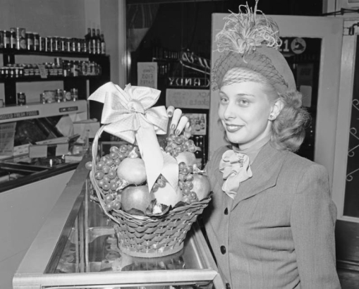 A woman poses with a basket of apples and grapes at the York Food Mart at 2021 East Colfax Avenue in Denver, Colorado. She wears a hat with feathers and a veil.
