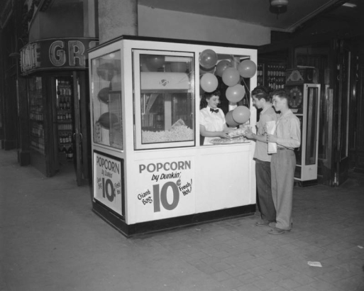 A teenaged boy and girl purchase popcorn at a stand festooned with balloons in (probably) Denver, Colorado. A sign reads: "Popcorn by Dunkin' 10 cents a Bag," a scale and shelves of liquor bottles are in a nearby concession; a neon sign reads: "Louie Gr...."