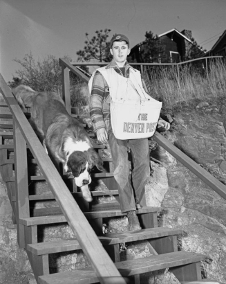 A teenage newspaper delivery boy, with a "Denver Post" newspaper bag, walks down stairs in Estes Park (Larimer County), Colorado; a Newfoundland dog is with him.