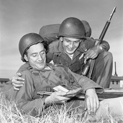 United States Army soldiers in fatigues look at photos in (probably) Korea. One holds a rifle, he has his arm around the other, who holds a wallet; both wear helmets and hold cigarettes.