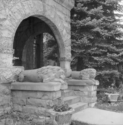 Partial exterior view of "Gargoyle" House, 302 West Pitkin Avenue, Pueblo, Colorado, shows the semi-elliptical arched porch of the rock-faced, stone, residence. Lion statues are atop rock-faced stone pedestals on either side of the entryway's steps.