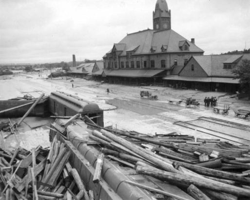 View of Union Depot in the aftermath of the 1921 flood in Pueblo, Colorado, shows debris and destruction. Piles of timber and beams are stacked against overturned railroad passenger cars. Mud and debris from the receded flood waters extends to the railroad station platform of Union Depot. Empty and loaded railroad baggage carts are next to a group of  men.