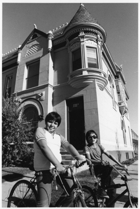 A boy and a girl (in sunglasses) pose on bicycles at the corner of 27th (Twenty Seventh) and Stout Streets in the Curtis Park Neighborhood of Denver, Colorado. A stone apartment building has corbeled brick, dentils, a corner turret, an arched window, and carved stone ornaments.