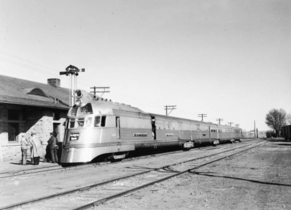 View of the Pioneer Zephyr  9900 running as Colorado and Southern Railway train 32 stopped at the railroad station in Longmont (Boulder County), Colorado. The streamlined art-deco style diesel locomotive and cars are constructed of stainless steel. The consist has a post office car, baggage car and passenger cars. Lettering on the train reads: "Burlington Route."