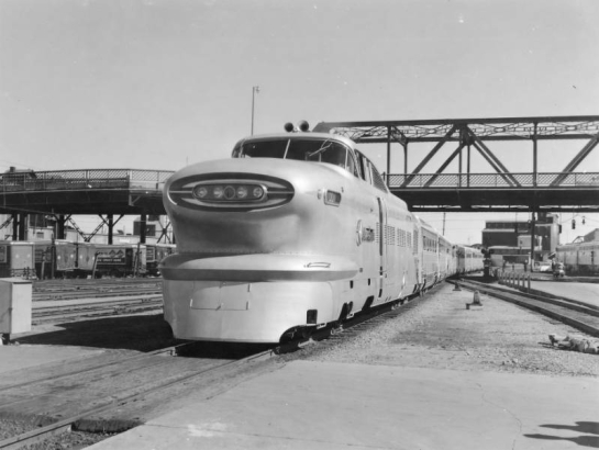 View of the General Motors experimental streamliner the Aerotrain led by a LWT12 locomotive at Union Station in Denver, Colorado.  The streamlined locomotive has four headlights. A steel viaduct and a flour mill are in the distance.