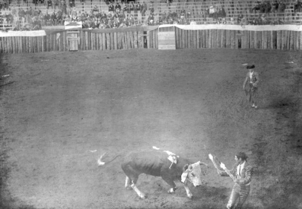 The bullfight on August 24 or 25, 1895, is taking place in the bullring built at the race track in Gillett, Colorado, for the occasion. Joe Wolfe was the organizer of the event which caused a scandal because of its cruelty to animals. Two of the Mexican bullfighters confront the bull; the matador holds a cape and stands at a distance, and the banderillo holds two banderillas directly in front of the pawing bull, who is already wounded. Spectators are in the stands behind the wood fence.