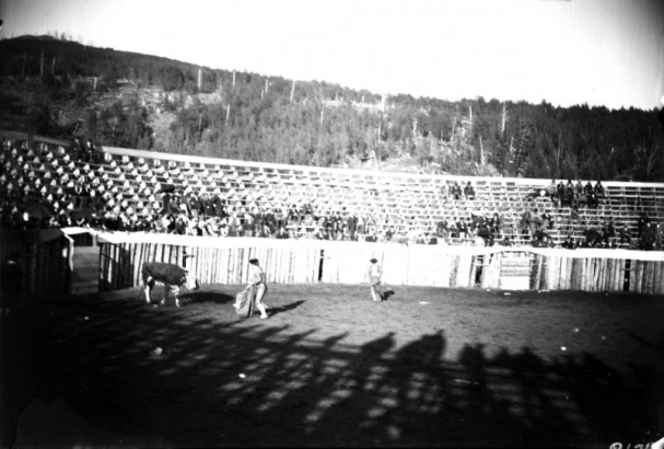 The bullfight on August 24 or 25, 1895, (the third day planned was cancelled) takes place in the bullring built at the race track in Gillett, Colorado, for the occasion. Joe Wolfe was the organizer of the event. The bull is coming out of the gate and the Mexican matador, probably  Jose Marrero, waves his cape; another bullfighter is in the ring. Spectators are in the stands behind the wood fence.