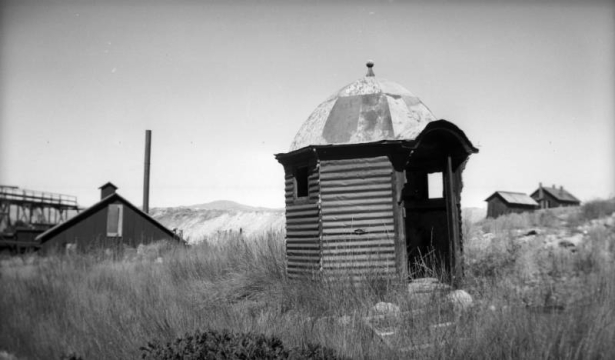 View of Finntown mining district's dilapidated structures in Stray Horse Gulch, east of Leadville (Lake County), Colorado. A small six-sided structure, possibly an outhouse, sits on a grassy slope in the foreground and is made of corrugated metal siding and has a faceted roof with finial ball, and a bracketed arched doorway. There are wood frame structures in the background and a tall smokestack on the left.