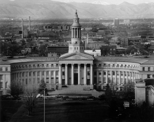 View of the Denver City and County Building at 1437 Bannock Street, in downtown Denver, Colorado; classic columns and entablature flank a pedimented entrance. A cupola / clock tower tops the curved facade; buildings, Front Range mountains and Mount Evans are in the distance.
