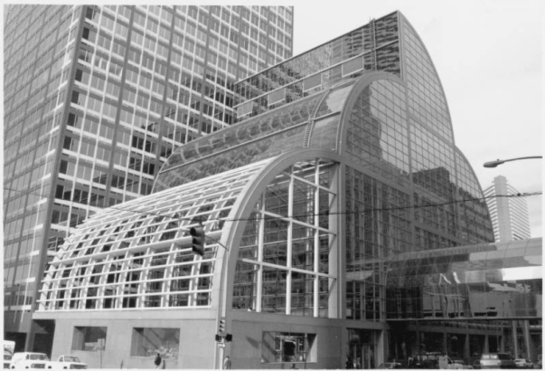 View of the United Bank Building at Wells Fargo Center, 1700 Lincoln Street in downtown Denver, Colorado. The glass atrium has a roofline of stepped curves; an elevated pedestrian walkway spans the street. Mile High Center (I. M. Pei, architect, 1956) is nearby.