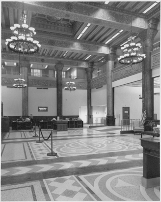 Interior view of the Women's Bank lobby at 821 17th (Seventeenth) Street in the central business district of Denver, Colorado. The large room has desks near a "Financial Sales & Services" sign, cast metal friezes beneath a wrought iron railing on the second floor loggia, and ornate chandeliers. The mastic or tile floor has a geometric pattern.