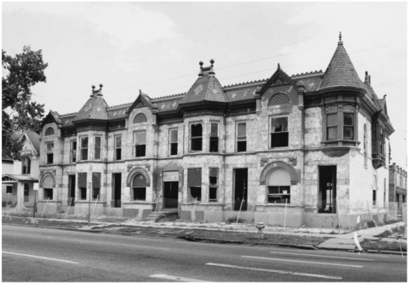 View of a two story stone row house at 2700 to 2714 Stout Street in the Five Points neighborhood of Denver, Colorado. The dilapidated house has High Victorian Gothic elements that include a corner turret, finials and a mansard roof. Shows missing doors and windows. A fence surrounds the property.