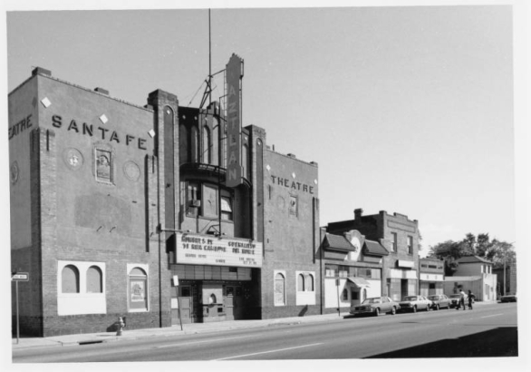 View of the Aztlan Theatre at 974 Santa Fe Drive in the Lincoln Park neighborhood of Denver, Colorado. The brick building has Art Deco elements, a stepped front and low relief designs. Signs read "Aztlan" and "Santa Fe Theatre." The marquee reads "Hombres De Ti[e]rra Caliente," "Guerrillero Del Norte," "Gerado Reyes," "Karate," and "Live Boxing, Oct. 19 Fri." Shows shops and commercial facilities beside the theater. Automobiles are parked along the street.