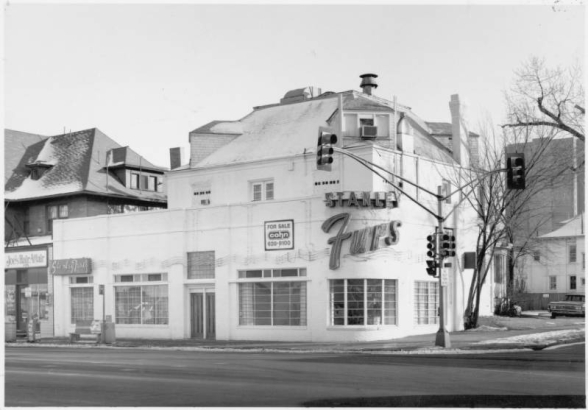 View of a business building at Colfax Avenue and Franklin Street in the Capitol Hill Neighborhood of Denver, Colorado. A storefront with a curved corner has been built onto a residence; a neon sign reads: "Stanley Furs." "Joe's Hair Affair" is next door.