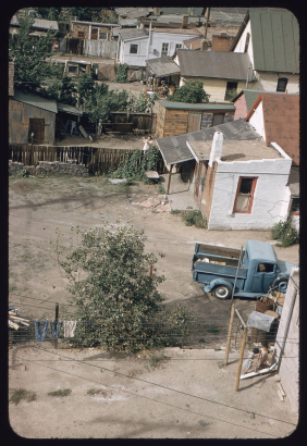 View of a pickup truck parked in a dirt backyard probably in the Sun Valley neighborhood, Denver, Colorado. Shows stuccoed brick houses, wooden fences and sheds and garages.