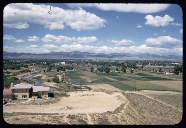 View of Arvada, Colorado, from Inspiration Point near 50th Avenue and Gray Street. Shows Clear Creek, the Wadsworth Drive-In Theater, the Arvada water tower, a two story stuccoed house, agricultural fields, and a bridge. Front Range mountains are in the distance.