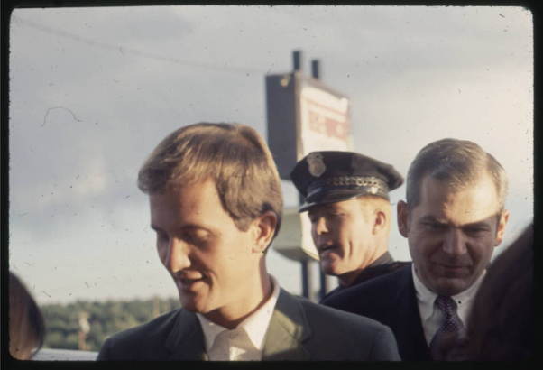 Pat Boone, a singer and actor, poses with an unidentified man, probably in Denver, Colorado.  A Colorado State trooper is nearby.
