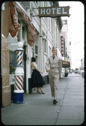 A young soldier in uniform walks down a street in the central Business District in Denver, Colorado. Shows a barber shop, a barber pole, and a sign for a drugstore. A woman leans against the wall of a hotel. She wears a blouse, skirt and high heels and carries a square plastic acrylic purse.