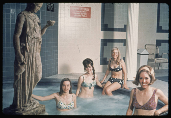 Women in two piece bathing suits pose on or in a jacuzzi type mineral bath in the club house of the Green Mountain Townhouses in Lakewood (Jefferson County), Colorado. A reproduction of a classical style statue of a woman is beside the pool. A sign reads: "Hydro-Swirl Mineral Pool 3 to 5 Minutes in This Pool is Sufficient." The walls are tiled. Shows a trash container and an aluminum and plastic chair.