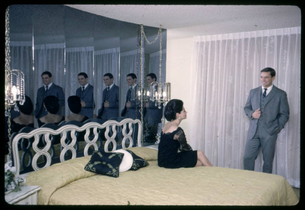 A man and a woman pose in a bedroom of a show unit at the Green Mountain Townhouses in Lakewood, Colorado. The room has a Queen sized bed with mirrors behind the headboard. Chandeliers that hang from chains hang over bedside tables. The woman wears a dark dress, has her hair pulled back in chignon and sits on the bed. The man wears a suit and tie and stands in front of a glass door covered with curtains.