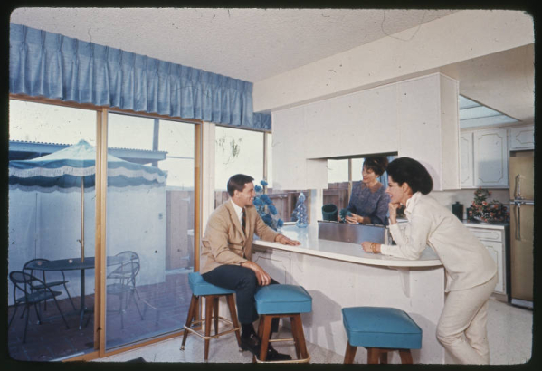 View of the kitchen area and breakfast bar in a unit at the Green Mountain Townhouses in Lakewood, Colorado. A glass patio door looks out on a brick patio with a table, an umbrella and chairs. A man sits at the breakfast bar on a stool. Women stand near the breakfast bar and a stove. Shows a refrigerator and cupboards. A valance is over the door.