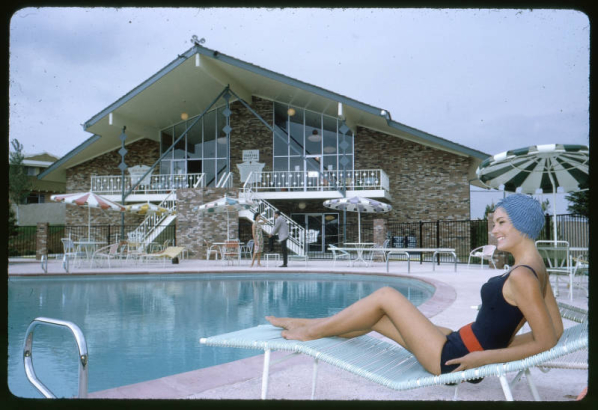 View of the clubhouse and swimming pool at the Green Mountain Townhouses in Lakewood, Colorado. The modern brick glass and steel building has an irregularly shaped pitch roof, wall to ceiling glass windows, balconies and external staircases. A woman sits in a chaise lounge near the pool. Shows outdoor tables, chairs and aluminum umbrellas.