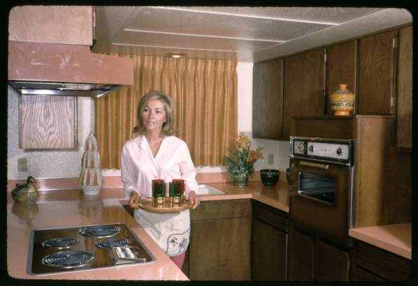 A woman stands in the kitchen of a model town home at the Green Mountain Townhouses in Lakewood, Colorado. She holds a tray of glasses and wears a white shirt, pink slacks, and an apron. Shows a built-in, wood grained, gas stove inset into a pink counter, an exhaust hood, a bronzed brown, built-in oven inset into a wooden enclosure and wood kitchen cabinets. Vases, artificial flowers and nic-nacs decorate the counters.