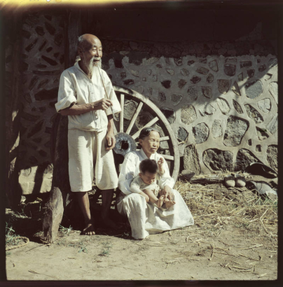 An elderly Korean man, his wife and a young boy pose near a stone and cement building in South Korea. The man wears linen knee length pants (paji) and a loose shirt with 3/4 length sleeves. He is balding and has a beard and long mustache. He smokes a long, bamboo and metal traditional Korean pipe. The woman sits beside the man and wears a white linen dress (hanbock). The child wears a sweater.