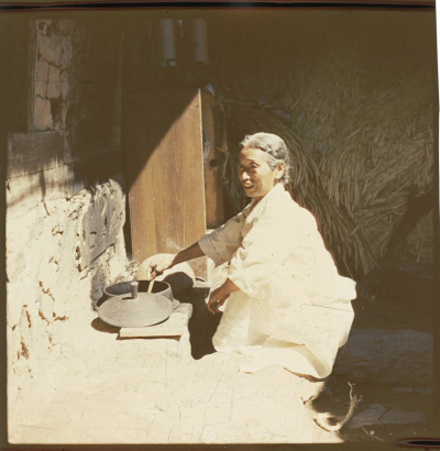 An elderly Korean woman kneels beside an outdoor hearth and stirs an iron pot in South Korea. The lid of the pot is beside the pot. The woman wears a loose fitting white linen traditional dress (hanbock). She wears her gray hair pulled back.