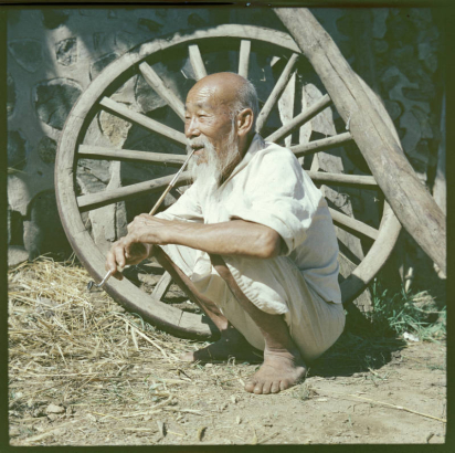 An elderly Korean man squats near a large wagon wheel that leans against a stone and cement wall in South Korea. He wears linen knee length pants (paji) and a loose shirt with 3/4 length sleeves. He is balding and has a beard and long mustache. He smokes a long, bamboo and metal traditional Korean pipe.