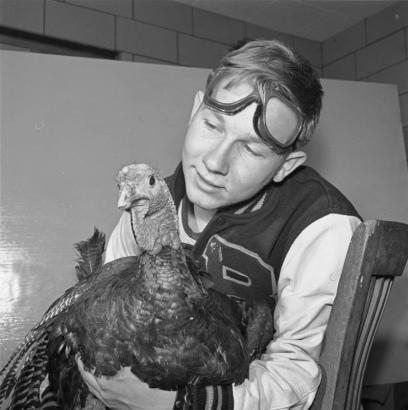 A teenage Red Cross volunteer poses with a live turkey, probably in Denver, Colorado. The boy wears a team jacket emblazoned with the letter "B." He has a pair of goggles on his head.