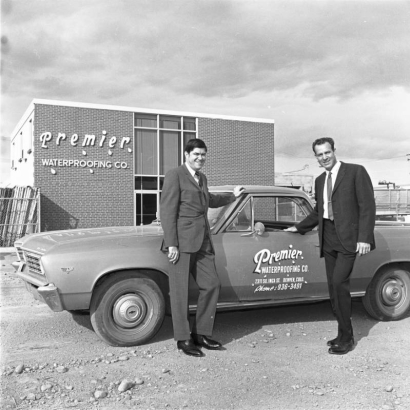 Two unidentified men in suits pose near a Chevrolet El Camino in front of Premier Waterproofing Company located at 2311 South Inca Street in the Overland neighborhood, Denver, Colorado. The brick building has a two story front glass and steel front. Lettering on the building reads: "Premier Waterproofing Co." Lettering on the truck reads: "Premier Waterproofing Co. 2311 So. Inca St. Denver, Colo. Phone: 936-3491."
