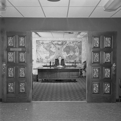 Interior view of probably the executive offices of the Acme Upholstery Supply Co., 1333 Wazee Street in the Auraria neighborhood, Denver, Colorado. Shows the door to an office flanked with wooden doors decorated with panels with stamped brass insets. A man sits at a desk, a large decorative world map is on the wall behind him.