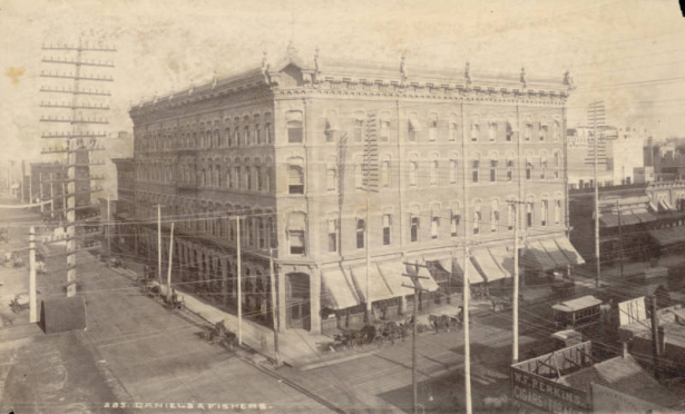 View of the corner, four-story Daniels & Fisher building at 16th (Sixteenth) and Lawrence Streets in the Central Business District in Denver, Colorado. Windows have hood molding and awnings and the flat roof line has decorative brackets and finials. Shows horse drawn carriages in the street, street car tracks on 16th Street and probably telephone poles. A sign reads: "W.F. Perkins, Cigars [and] Tobacco."