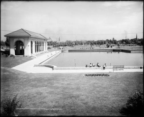 View of covered pavilion with archways and pond, Sunken Gardens, Denver, Colorado; shows background view of Denver skyline including Gas & Electric building, Daniels and Fisher tower, Tramway building, Arapahoe County Courthouse, and Evans Chapel (steeple or spire on far right). Boys sit on cement sides of raised pond or lake. View includes wrought-iron park benches, landscaping, flower gardens, water sprinklers, and 11th (Eleventh) Avenue viaduct over Cherry Creek.
