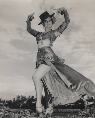 Louise Campa, Lucille Campa, a well known dancer and teacher of Mexican, Spanish classical dance and flamenco, poses in a classical style Spanish dance costume. She wears a short blouse with long sleeves, has a bare midriff and has a short skirt with a long train. She wears a Spanish style wide brimmed hat and holds castanets. Louise Campa was the wife of Professor Arthur L. Campa, Sr., and author, renowned scholar, historian and professor of cultural anthropology.