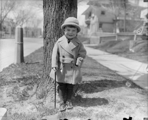 A boy poses outdoors with a cane possibly in Denver, Colorado. He wears a coat and bowler.