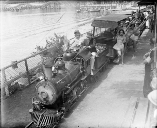 A group of women pose with the conductor on a miniature train at Lakeside Amusement Park in Denver, Colorado. Lake Rhoda and a roller coaster are nearby.