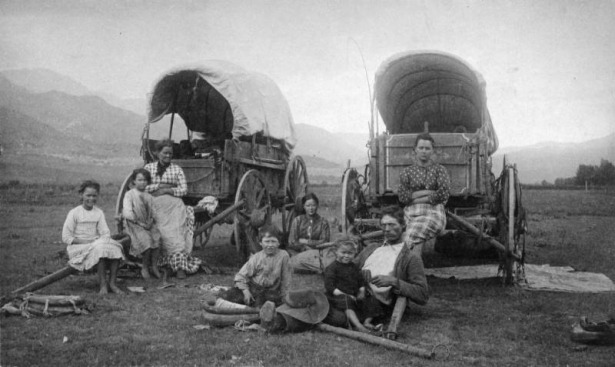 Weary pioneers rest in front of two Conestoga wagons on a plain with mountains in the background. Family includes an adult male, 3 adult females, and 4 children.