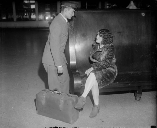 A man in a porter uniform talks to a woman seated on a wooden bench at Union Station, in Denver, Colorado. The woman wears a fur coat and has a suitcase.