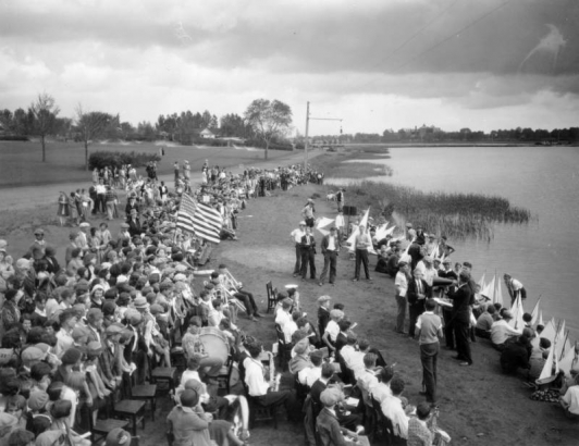 Young boys with toy sailboats gather at the shore of Sloans Lake at Sloans Lake Park in the Sloan Lake neighborhood of Denver, Colorado. The school band is seated with instruments in hand. Students and observers crowd near the shore. Three African-American (Black) students are in the crowd.