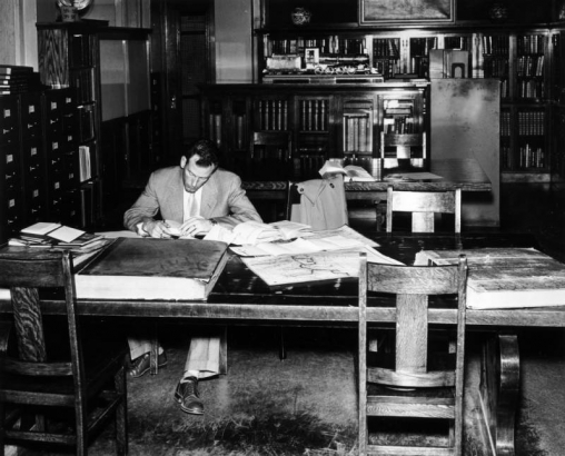 A man reads at a table in the Western History department of the Denver Public Library at Colfax Avenue and Bannock Street in the Civic Center neighborhood of Denver, Colorado. Maps and large, bound books are on the table. Bookcases, filing cabinets, and model of a Colorado Midland locomotive and coal car are in the room.