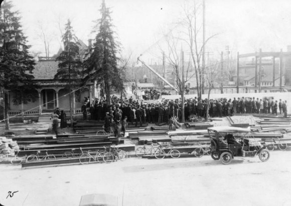 People crowd the Denver Public Library (Carnegie) construction site by a crane to watch the cornerstone ceremony in the Civic Center neighborhood of Denver, Colorado. An automobile and bicycles are parked; the Burton house with a covered porch is to the side.