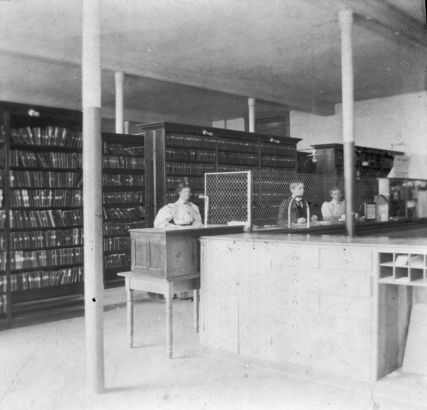 Interior view of the Mercantile Library in Denver, Colorado; shows a man and woman working behind a counter by bookshelves.