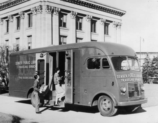 View of the Denver Public Library bookmobile in Denver, Colorado; lettering reads: "Denver Public Library - Traveling Branch." A man and woman board the vehicle; the Carnegie Building of the Denver Public Library in Civic Center Park is in the background.