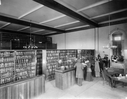 Interior view of the Denver Public Library at East Denver High School in Denver, Colorado; shows people at a counter, bookshelves, a coffered ceiling and hanging lamps. John Cotton Dana is behind the desk.