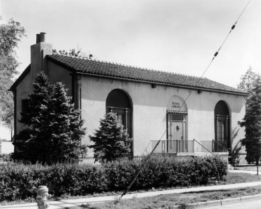 View of the Elyria branch of the Denver Public Library located on 47th Avenue and High Street in the Elyria-Swansea neighborhood of Denver, Colorado. The spanish style building was designed by architect H. J. Manning; it features a tile roof, arched windows, iron lamps, and a projecting doorway. The Elyria branch was closed in 1952.