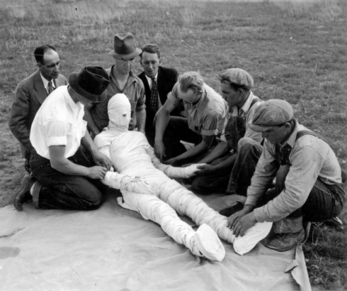 Men engage in first aid instruction in Denver, Colorado; the "victim" is wrapped in bandages.